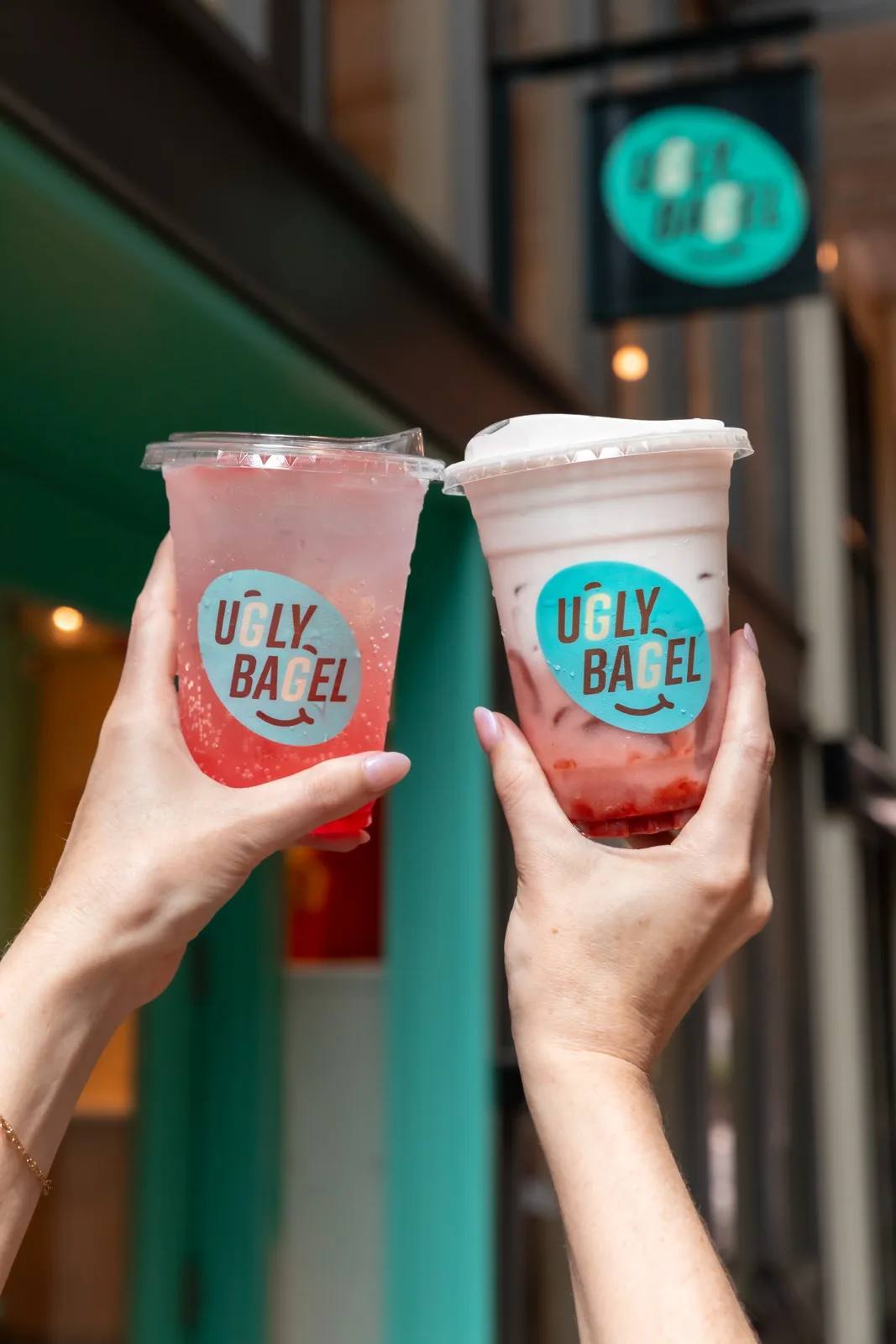 Two hands toasting pink Ugly Bagel drinks